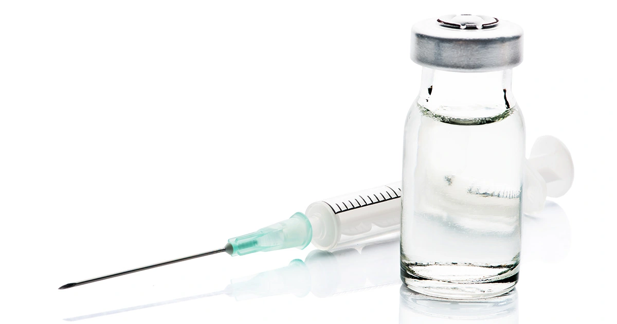 A close-up image of a syringe filled with liquid lying next to a vial containing a clear solution, both placed on a reflective white surface