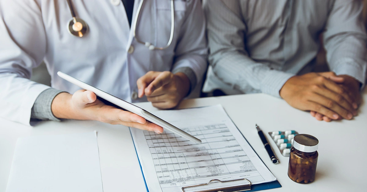 A close-up image of a doctor holding a tablet and discussing it with a patient. The doctor and patient are seated at a desk with medical forms, a pen, and a bottle of medication on the table