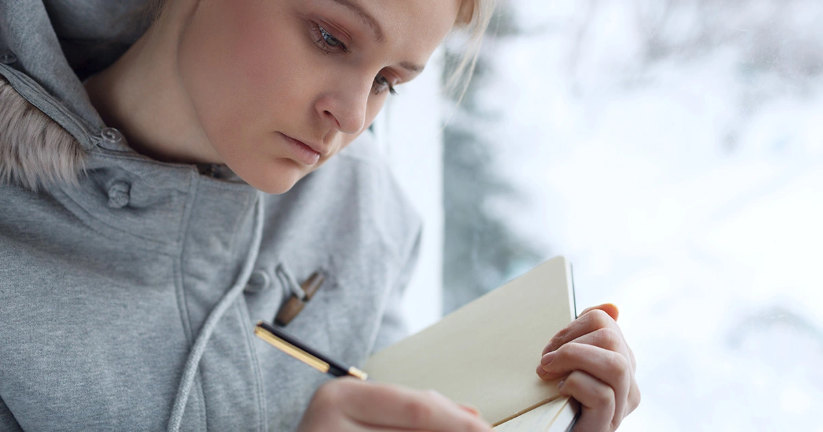 A woman in a grey hoodie is sitting by a window on a cold day, writing in a notebook.