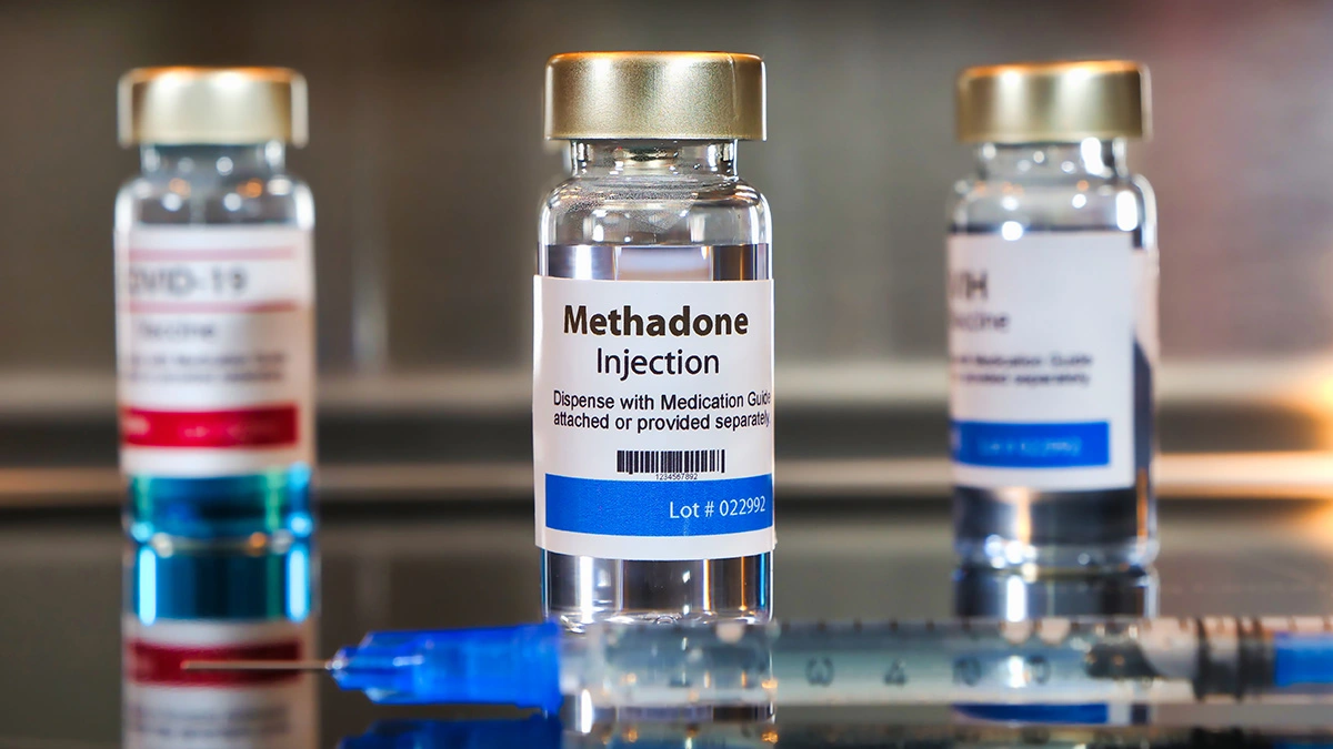 "A vial labeled 'Methadone Injection' is prominently displayed on a reflective surface, accompanied by a syringe in the foreground.